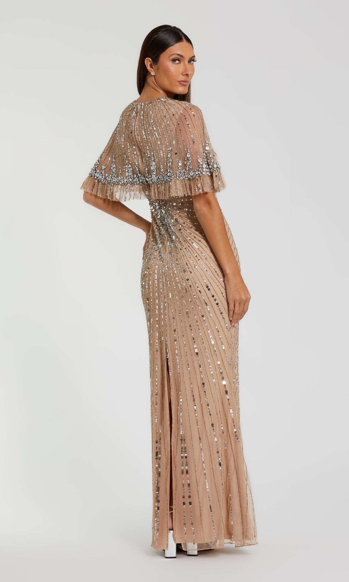 A woman stands looking over her shoulder in the Mac Duggal 23044 Capelet Long Beaded Formal Dress, a floor-length nude gown adorned with silver sequins and featuring a cape detail.